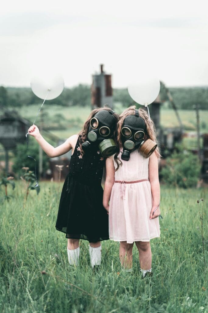 Two children in dresses and gas masks hold balloons in an overgrown field.