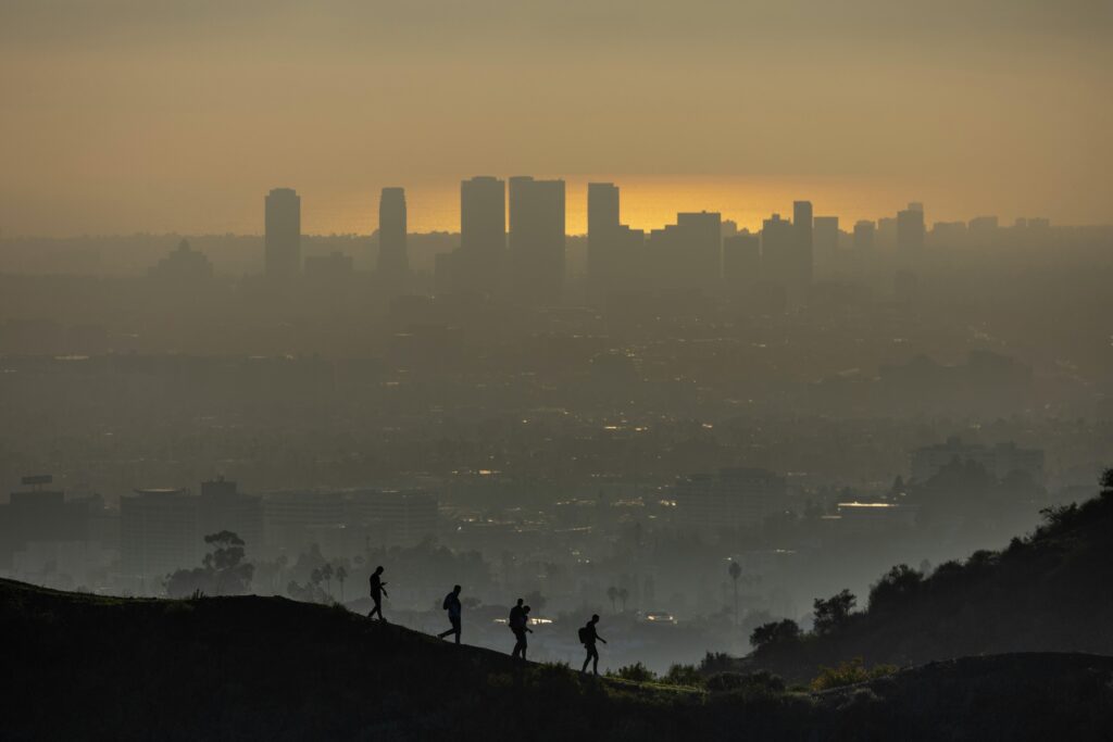 Silhouetted hikers traverse a hill, overlooking the smoggy Los Angeles skyline at sunset.